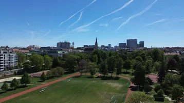 Luftaufnahme eines Stadtparks mit Sportanlagen und umliegenden Gebäuden unter blauem Himmel.