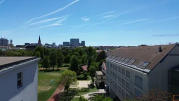Panoramablick über die Stadt mit grünen Parkanlagen, Wohngebäuden und einer fernen Skyline unter blauem Himmel.