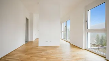 Helles, leeres Zimmer mit Holzboden und großen Fenstern, die einen Blick ins Grüne bieten.