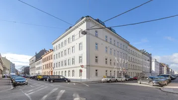 Eckgebäude in urbaner Umgebung mit klassischer Architektur und vielen Fenstern an einem sonnigen Tag.