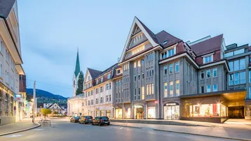 Abendliche Ansicht einer belebten Straße mit historischen Gebäuden, Geschäften und einem Kirchturm im Hintergrund.