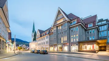 Gebäudeansicht bei Dämmerung mit beleuchteten Fenstern und Geschäften an einer städtischen Straße.