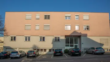 Gebäudeansicht mit Parkplätzen davor und Fenstern mit Rollläden unter blauem Himmel.