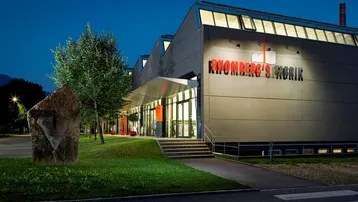 Modernes Gebäude mit beleuchteter Fassade und Eingangsbereich bei Dämmerung, umgeben von Grünflächen.