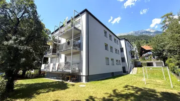 Mehrfamilienhaus mit Balkonen, umgeben von Grünflächen und mit Bergblick im Hintergrund.
