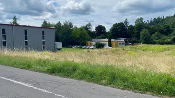 Großes, unbebautes Grundstück mit angrenzendem Gewerbegebäude und bewaldetem Hügel im Hintergrund.