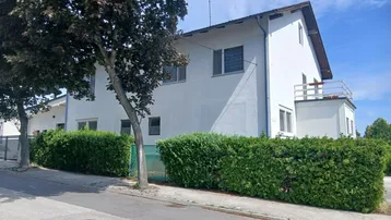Weißes Wohnhaus mit grüner Hecke und Bäumen an einer Straße unter blauem Himmel.
