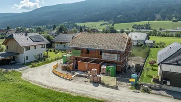 Baustelle eines Einfamilienhauses im Rohbau mit Dachstuhl, umgeben von grüner Landschaft.