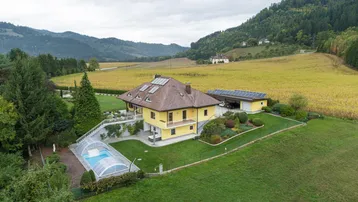 Luftaufnahme eines gelben Hauses mit Pool, umgeben von grünen Wiesen und bewaldeten Hügeln.