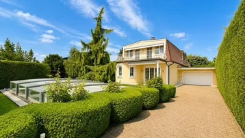 Gelbe Villa mit gepflasterter Auffahrt, Garage und überdachtem Pool im gepflegten Garten.