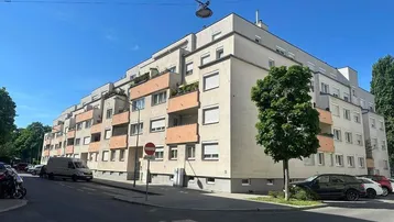 Mehrstöckiges Wohngebäude mit Balkonen und Rollläden an einem sonnigen Tag in städtischer Umgebung.