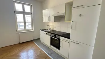 Helle Küche mit weißen Einbauschränken, schwarzer Arbeitsplatte und integrierten Geräten sowie Parkettboden.