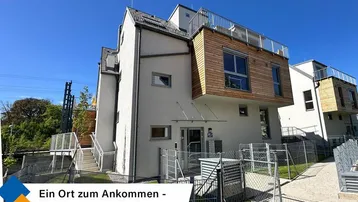 Modernes Gebäude mit weißer Fassade, Holzelementen und großen Fenstern unter blauem Himmel.