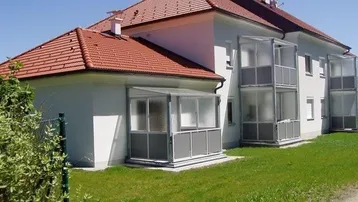 Außenansicht eines Wohnhauses mit rotem Ziegeldach, weißer Fassade und verglasten Balkonen.