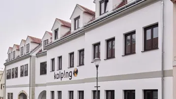 Weißes Gebäude mit braunen Fenstern und Dachgauben an einer nassen Straße.