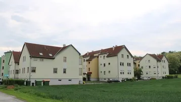 Reihenhausanlage mit hellen Fassaden und roten Ziegeldächern, umgeben von grünen Feldern unter bewölktem Himmel.
