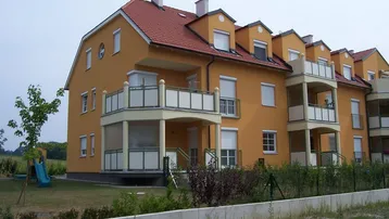 Mehrfamilienhaus mit gelber Fassade, rotem Ziegeldach und Balkonen, umgeben von Grünflächen und einem kleinen Spielplatz.