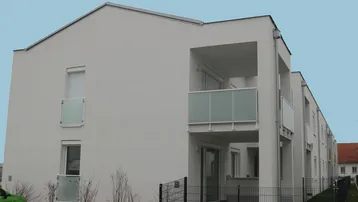 Modernes Mehrfamilienhaus mit weißer Fassade und verglasten Balkonen, umgeben von einer kleinen Grünfläche.