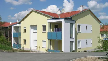 Modernes Mehrfamilienhaus mit hellgelber Fassade, roten Ziegeldächern und Balkonen mit blauen Geländern unter blauem Himmel.