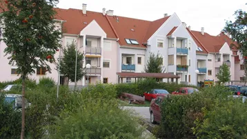 Mehrfamilienhaus mit farbiger Fassade, roten Ziegeldächern und Balkonen, umgeben von viel Grün.