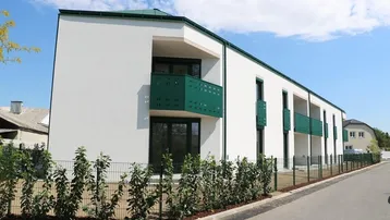Modernes Neubaugebäude mit weißer Fassade, grünen Balkonen und flachem Dach in einer Wohngegend.