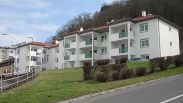 Weiße Mehrfamilienhäuser mit grünen Balkonen und Fenstern an einem Hang gelegen, umgeben von Natur.