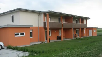 Modernes Mehrfamilienhaus mit orangefarbener Fassade, Balkonen und Grünflächen in ländlicher Umgebung.