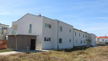 Modernes Mehrfamilienhaus mit heller Fassade, Balkonen und einem angrenzenden Holzschuppen in ländlicher Umgebung.