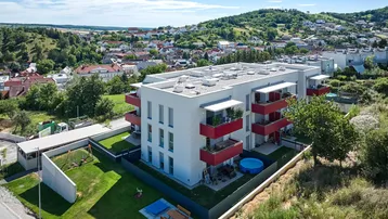 Modernes Mehrfamilienhaus mit roten Balkonen, umgeben von viel Grün und einem Spielplatz, in ländlicher Umgebung.