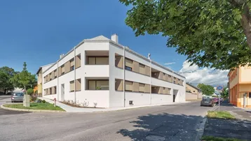 Modernes weißes Mehrfamilienhaus an einer Straßenecke mit großen Fenstern, Außenjalousien und kleinen Grünflächen.