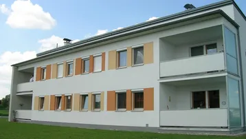 Modernes Mehrfamilienhaus mit weißer Fassade, farbigen Fensterakzenten und Balkonen, umgeben von einer grünen Rasenfläche.