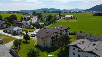 Luftaufnahme des Mehrfamilienhauses in ländlicher Umgebung mit weiten Feldern und Bergen im Hintergrund.
