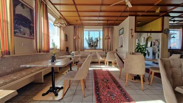 Geräumiger Speisesaal mit Holzkassettendecke, Fliesenboden und mehreren Tischen, hell durch große Fenster.