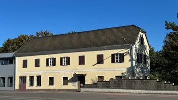 Gelbes Wohnhaus mit braunem Dach und Fensterläden unter blauem Himmel.
