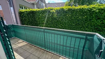 Balkon mit Fliesenboden und grüner Sichtschutzwand, umgeben von einer hohen Hecke.