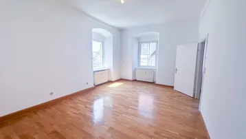 Helles, leeres Zimmer mit Parkettboden, weißen Wänden und zwei Fenstern, die viel Tageslicht hereinlassen.