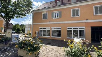 Gebäudeansicht mit orangefarbener Fassade, Fenstern mit Fensterläden und Blumenkübeln im Vordergrund.