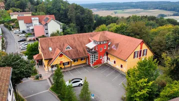 Luftaufnahme eines großen Gebäudes mit rotem Dach und gelber Fassade, umgeben von Bäumen und einem Parkplatz.