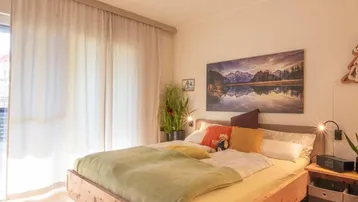Helles Schlafzimmer mit Holzbett, großem Fenster und Bergbild an der Wand.