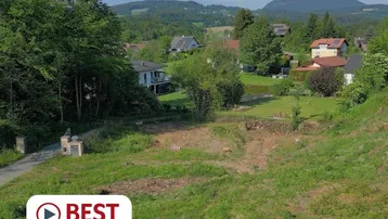 Blick auf ein unbebautes Grundstück mit umliegenden Häusern, Bäumen und Bergen im Hintergrund.