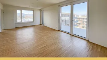Helles, leeres Wohnzimmer mit Parkettboden und großen Fenstern, die Zugang zu einem Balkon bieten.
