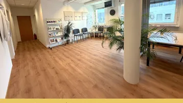 Heller Büroraum mit Laminatboden, großen Fenstern und einer Wartezone mit Pflanzen.