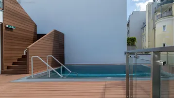Moderner Außenpool auf einer Holzterrasse mit Treppenzugang und umliegenden Gebäuden.
