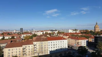 Panoramablick über eine Stadt mit vielen Gebäuden, roten Dächern und einem markanten Turm unter blauem Himmel.