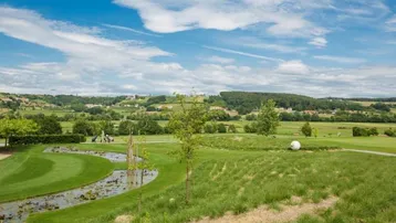 Weitläufige Landschaft mit einem Golfplatz, einem kleinen Bach und einem Dorf in der Ferne unter blauem Himmel.
