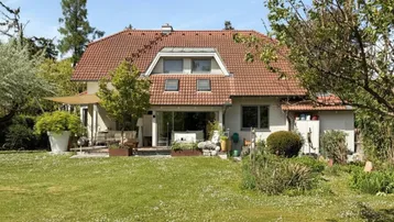 Hausrückseite mit großer Rasenfläche, Terrasse und üppiger Bepflanzung unter blauem Himmel.