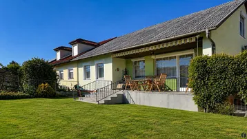 Hausfassade mit großer Rasenfläche und einer möblierten Terrasse unter einer Markise.
