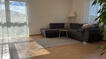 Lichtdurchflutetes Wohnzimmer mit gemütlichem Ecksofa, Holzboden und Blick auf die umliegende Landschaft.