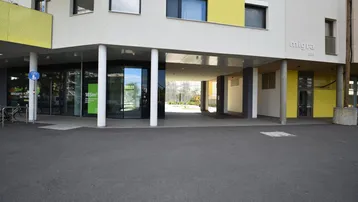 Modernes Gebäude mit Geschäften im Erdgeschoss und Wohnungen darüber, gekennzeichnet durch gelbe Akzente und eine offene Passage.