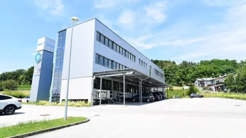 Modernes Bürogebäude mit heller Fassade, großen Fenstern und Parkplätzen im Freien unter blauem Himmel.